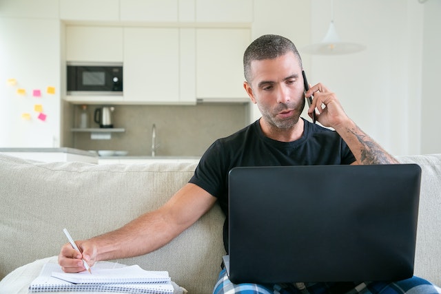 seorang pria sedang menatap layar laptop sambil melakukan panggilan via handphone